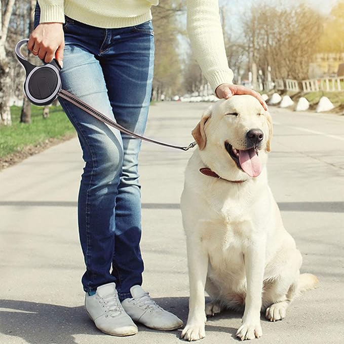 Une personne en jean et pull blanc se tient à l'extérieur par une journée ensoleillée, tenant le MaxLeash™ - Noir tout en caressant doucement un heureux Labrador jaune assis sur le trottoir, avec des arbres visibles en arrière-plan.
