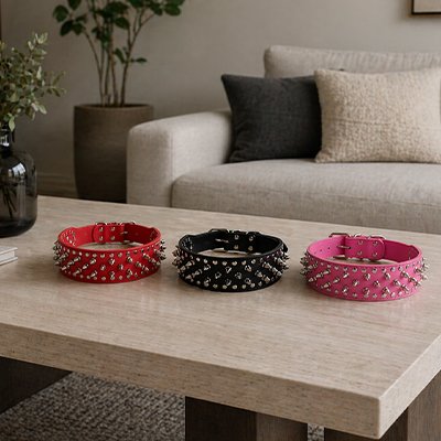 Trois colliers en cuir pour chien, rouge, noir et rose, ornés de pointes métalliques, posés sur une table dans un salon moderne.